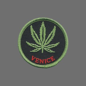May include: Circular embroidered patch with a green cannabis leaf on a black background. The word "VENICE" is embroidered in red. The patch is edged with green stitching.