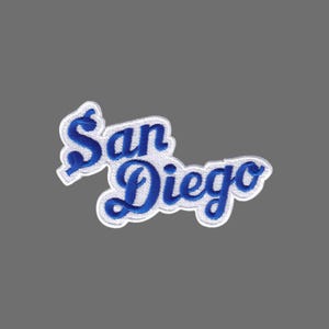 May include: Embroidered patch featuring the city name "San Diego" in a stylized, cursive font. The text is in a vibrant blue color, outlined with white stitching. The patch has a white border and is set against a gray background.