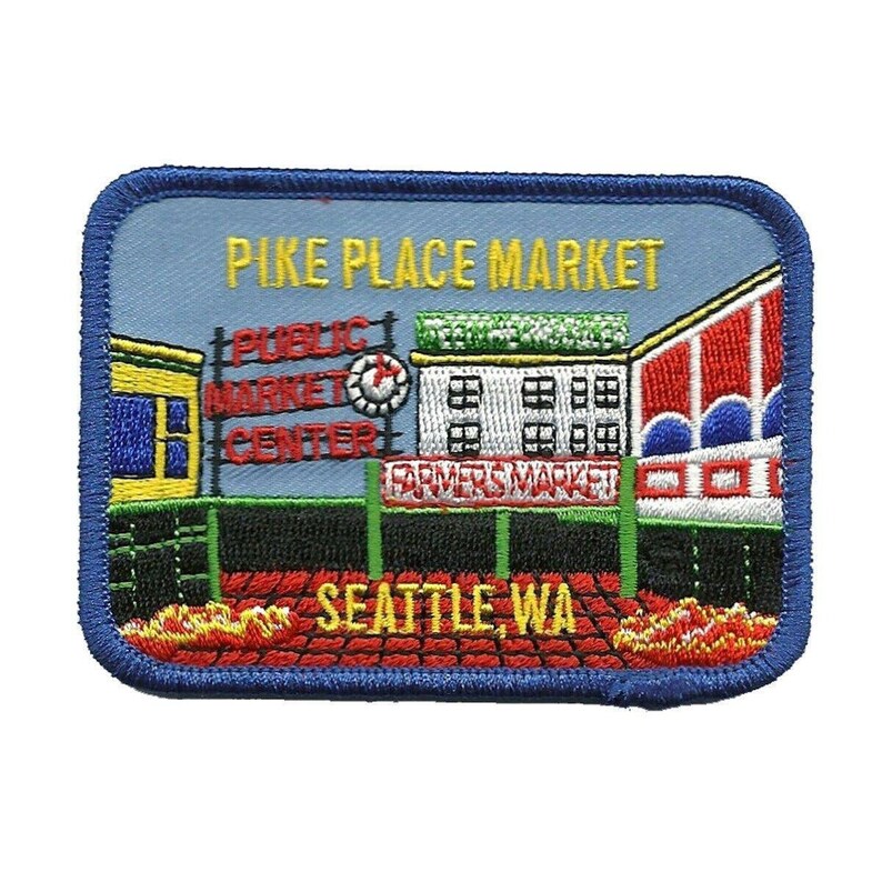 May include: Embroidered patch depicting Pike Place Market in Seattle, Washington. The patch features a blue background with a white building and red, white, and blue awnings. The text "Pike Place Market" is in yellow lettering. The text "Public Market Center" is in red lettering. The text "Farmers Market" is in green lettering. The text "Seattle, WA" is in red lettering.