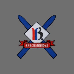 May include: Embroidered patch featuring the word "BRECKENRIDGE" on a red banner. The patch has a white diamond shape with the letters "1B" in red, blue, and black. Two blue crossed skis are behind the diamond.
