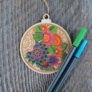 May include: A wooden bauble ornament shaped like a Christmas tree with a colourful floral design. The ornament is decorated with green, red, pink, blue and yellow flowers. Two markers, one green and one blue, are lying next to the ornament.