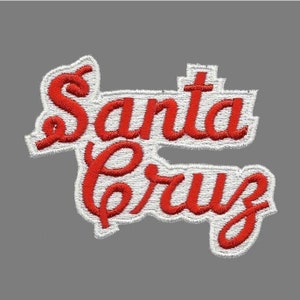 May include: Embroidered patch featuring the words "Santa Cruz" in red script lettering. The text is outlined in white stitching and set against a gray background. The patch is likely for clothing or accessories.