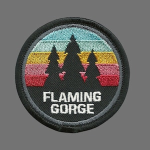 May include: A black and white embroidered patch with a retro sunset design featuring three pine trees and the text "FLAMING GORGE".