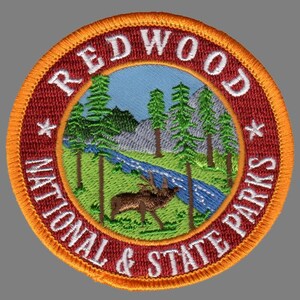 May include: Redwood National & State Parks embroidered patch with a red background, orange border, and a white star on each side of the text. The center of the patch features a green and brown landscape with a blue river, a brown elk, and green trees.