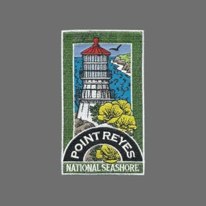 May include: Embroidered patch featuring a lighthouse with a red roof, set against a blue sky and ocean. The patch includes the text "POINT REYES NATIONAL SEASHORE" in a semi-circular design, with yellow flowers in the foreground.