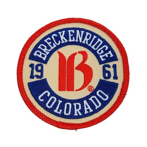 May include: A circular embroidered patch with a red "B" in the center, surrounded by a blue ring with the text "Breckenridge 19 61 Colorado" in white. The patch has a red outer border.