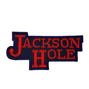 May include: A navy blue embroidered patch with the words "JACKSON HOLE" in red block letters. The patch has a unique shape with a blue border.