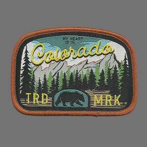 May include: Embroidered patch with a brown border, featuring a mountain landscape with a bear silhouette and the text "My heart is in Colorado" in yellow lettering. The text "TRD MRK" is also visible at the bottom.