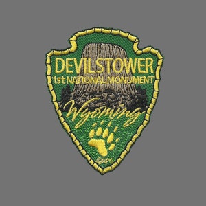 May include: Green and gold embroidered patch with the text "Devil's Tower 1st National Monument Wyoming" and a paw print.