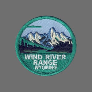 May include: Embroidered patch featuring a mountain range scene. The patch is circular with a teal border and a light blue sky background. The text "WIND RIVER RANGE WYOMING" is embroidered in white.