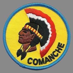 May include: A round, blue-trimmed patch with a yellow background. A cartoon image of a Native American man wearing a red, white, and black headdress. The text "COMANCHE" is written in black letters around the bottom of the patch.