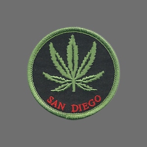 May include: Embroidered patch featuring a green cannabis leaf on a black background, encircled by green stitching. The text "SAN DIEGO" is stitched in red at the bottom. The patch is round.