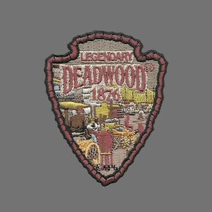 May include: A brown and tan embroidered patch shaped like an arrowhead with the text "LEGENDARY DEADWOOD 1876" in white and black lettering. The patch has a red border and a small brown label with the text "G.P.I."
