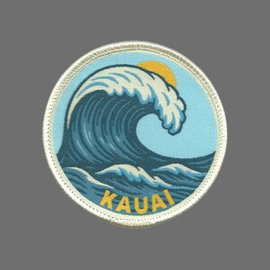 May include: Round patch featuring a stylized ocean wave in shades of blue and white, with a yellow sun in the background. The word "KAUAI" is printed in yellow at the bottom. The patch has a silver border.