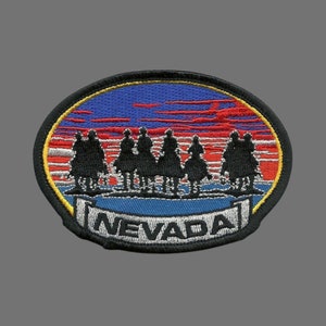 May include: An embroidered patch with a sunset scene and a group of riders silhouetted against the horizon. The patch is oval-shaped and has a blue and red background with a yellow border. The text "NEVADA" is written in a white banner at the bottom of the patch.