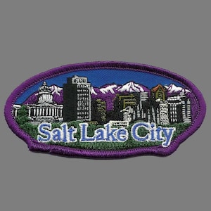 May include: Purple oval embroidered patch with a cityscape of Salt Lake City, Utah, including the Salt Lake Temple and mountains in the background. The text "Salt Lake City" is embroidered in blue on a green background.