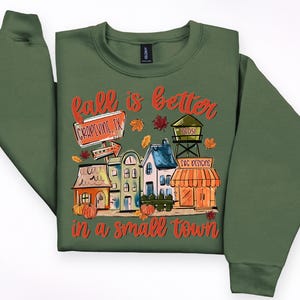May include: Green sweatshirt with a colorful graphic that reads "Fall is better in a small town." The graphic features a town scene with houses, pumpkins, and the text "Grapevine, TX."