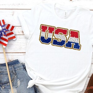 Puede incluir: Camiseta blanca con un gr&aacute;fico de "USA" en purpurina roja, blanca y azul.