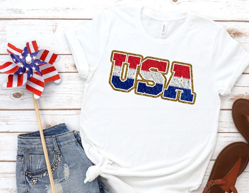 Puede incluir: Camiseta blanca con un gr&aacute;fico de "USA" en purpurina roja, blanca y azul.