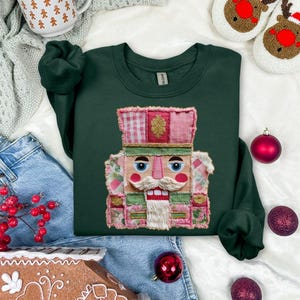 May include: A dark green sweatshirt featuring a colorful, patchwork-style nutcracker design. The nutcracker has a white mustache and beard, with pink, green, and red accents. The sweatshirt is surrounded by Christmas ornaments and holiday-themed items.