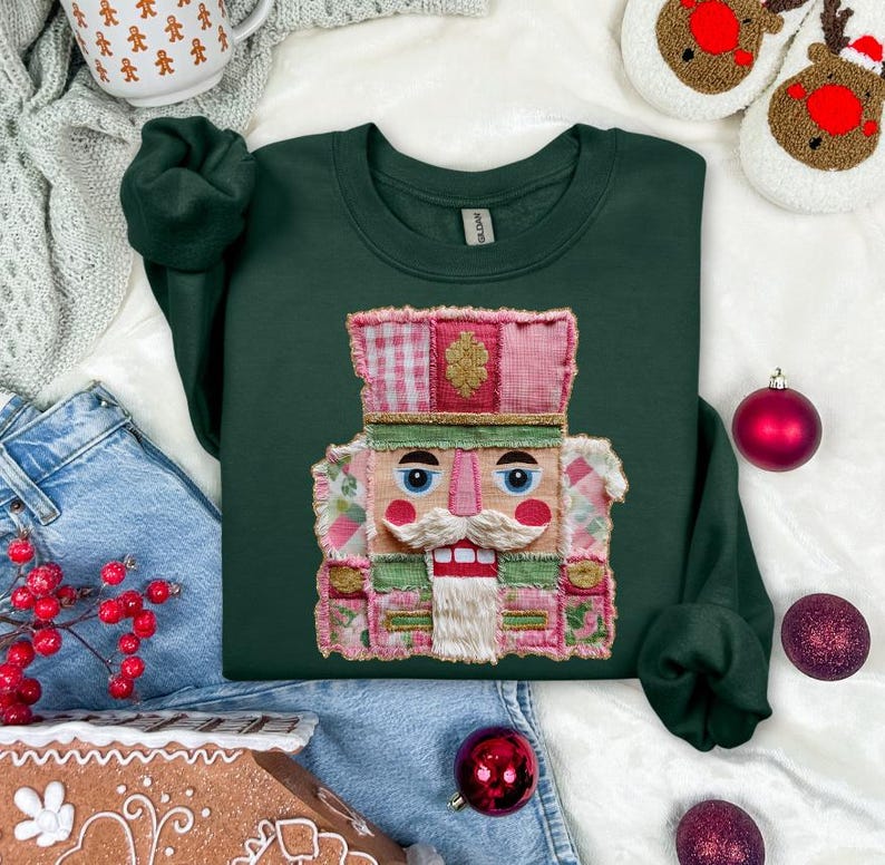 May include: A dark green sweatshirt featuring a colorful, patchwork-style nutcracker design. The nutcracker has a white mustache and beard, with pink, green, and red accents. The sweatshirt is surrounded by Christmas ornaments and holiday-themed items.