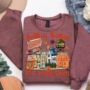 May include: Burgundy crewneck sweatshirt with a colorful graphic. The graphic reads "Fall is better in a small town" and features a town scene with pumpkins, leaves, and buildings. The sweatshirt is made of a soft material.