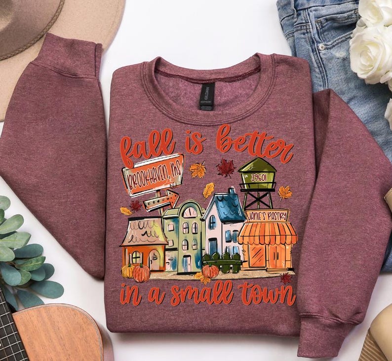 May include: Burgundy crewneck sweatshirt with a colorful graphic. The graphic reads "Fall is better in a small town" and features a town scene with pumpkins, leaves, and buildings. The sweatshirt is made of a soft material.