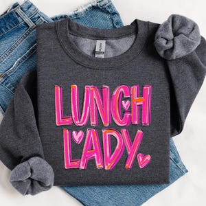 May include: A dark gray sweatshirt with the text "Lunch Lady" in pink and white lettering with pink hearts.