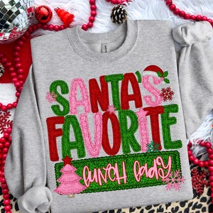 May include: A heather gray sweatshirt with the text "SANTA'S FAVORITE LUNCH LADY" in green, red, and pink. The design includes a Santa hat, snowflakes, and a pink Christmas tree. Festive apparel for the holiday season.