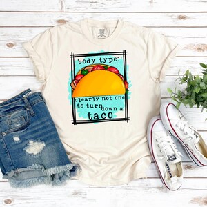 May include: A beige t-shirt with a graphic of a taco and the text "body type: clearly not one to turn down a taco".