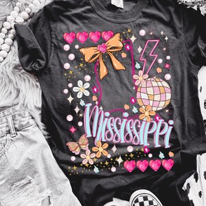 Puede incluir: Camiseta negra con un dise&ntilde;o gr&aacute;fico rosa, azul y naranja que presenta una bola de discoteca, un lazo y la palabra "Mississippi".