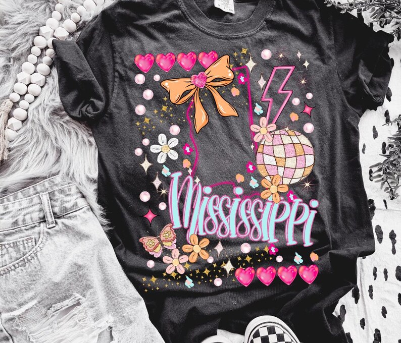 Puede incluir: Camiseta negra con un dise&ntilde;o gr&aacute;fico rosa, azul y naranja que presenta una bola de discoteca, un lazo y la palabra "Mississippi".