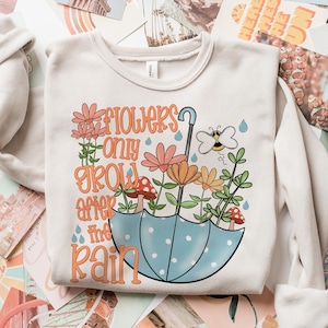 May include: A white sweatshirt featuring a graphic design of flowers, a bee, and an umbrella. The text on the sweatshirt reads "Flowers only grow after the rain."