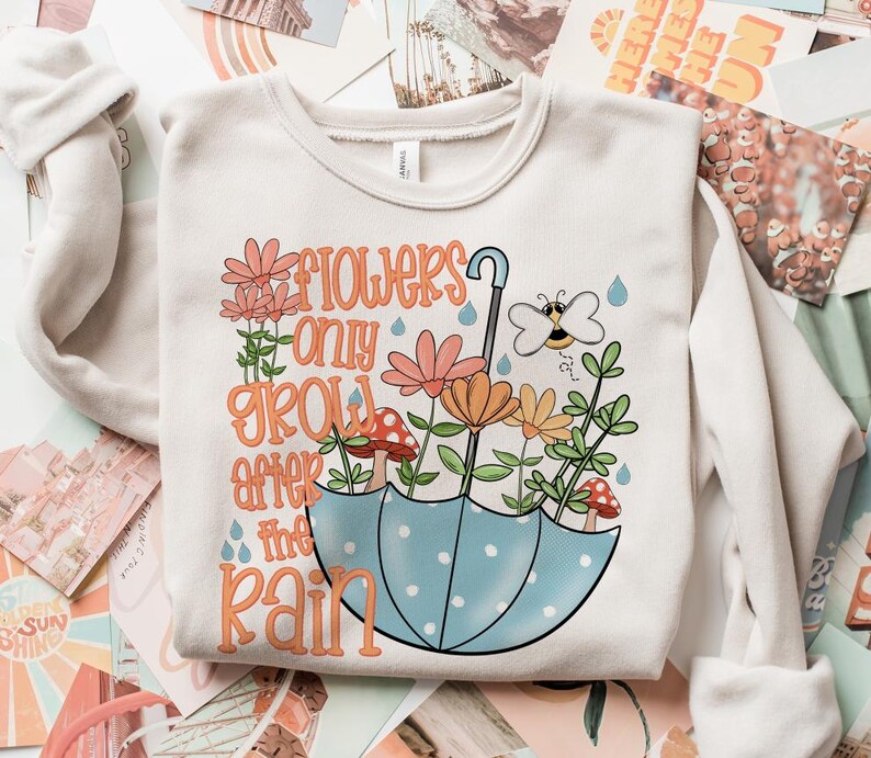May include: A white sweatshirt featuring a graphic design of flowers, a bee, and an umbrella. The text on the sweatshirt reads "Flowers only grow after the rain."