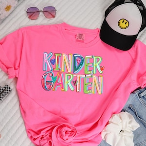 May include: A pink t-shirt with the word "KINDERGARTEN" printed in colorful letters with hearts. The letters are outlined in black and have a rainbow color scheme.