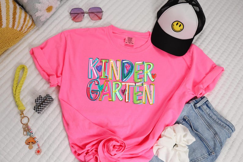 May include: A pink t-shirt with the word "KINDERGARTEN" printed in colorful letters with hearts. The letters are outlined in black and have a rainbow color scheme.