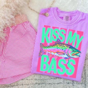 May include: A lilac Comfort Colors t-shirt with a neon green and pink graphic that says "KISS MY BASS" and features a colorful fish illustration. Beside the shirt is a pair of pink gingham shorts.