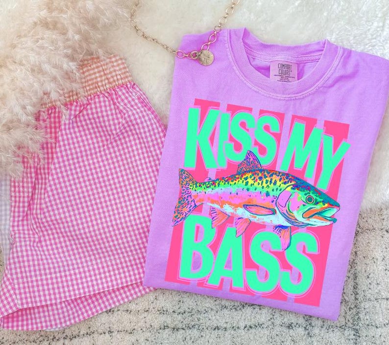 May include: A lilac Comfort Colors t-shirt with a neon green and pink graphic that says "KISS MY BASS" and features a colorful fish illustration. Beside the shirt is a pair of pink gingham shorts.