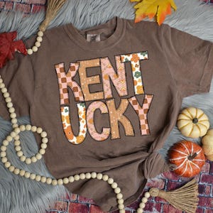 May include: A brown t-shirt with a patchwork design that reads "Kentucky" in a retro font. The shirt is on a gray fuzzy surface with fall-themed decorations.