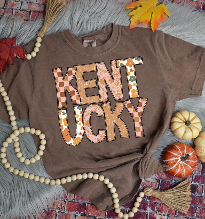 May include: A brown t-shirt with a patchwork design that reads "Kentucky" in a retro font. The shirt is on a gray fuzzy surface with fall-themed decorations.