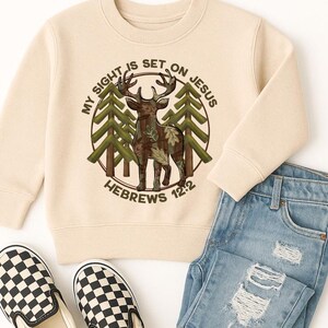 May include: A cream-colored sweatshirt featuring a circular graphic with a deer, trees, and the text "MY SIGHT IS SET ON JESUS" and "HEBREWS 12:2". The sweatshirt is paired with blue jeans and black and white checkered slip-on shoes.
