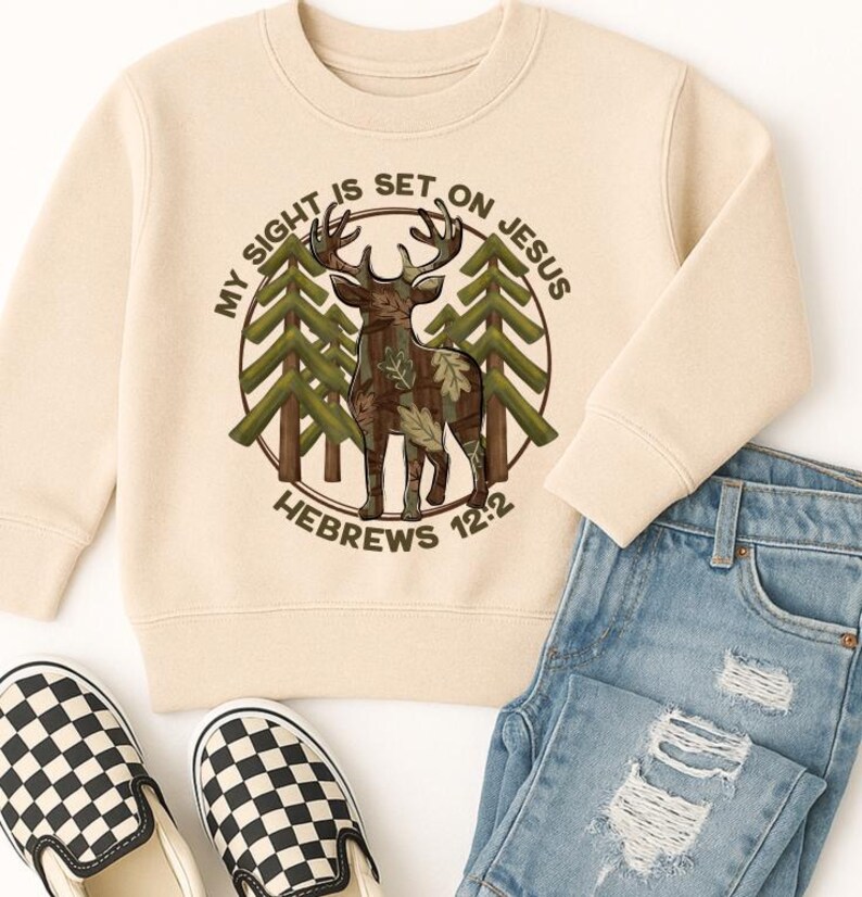 May include: A cream-colored sweatshirt featuring a circular graphic with a deer, trees, and the text "MY SIGHT IS SET ON JESUS" and "HEBREWS 12:2". The sweatshirt is paired with blue jeans and black and white checkered slip-on shoes.