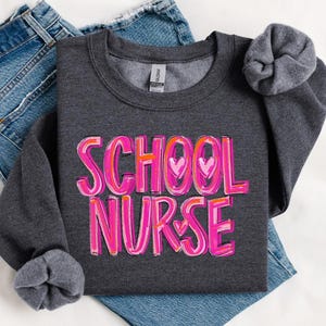 May include: A dark gray sweatshirt with the words "SCHOOL NURSE" in pink and orange lettering with hearts. The sweatshirt is folded and laid on a pair of blue jeans.