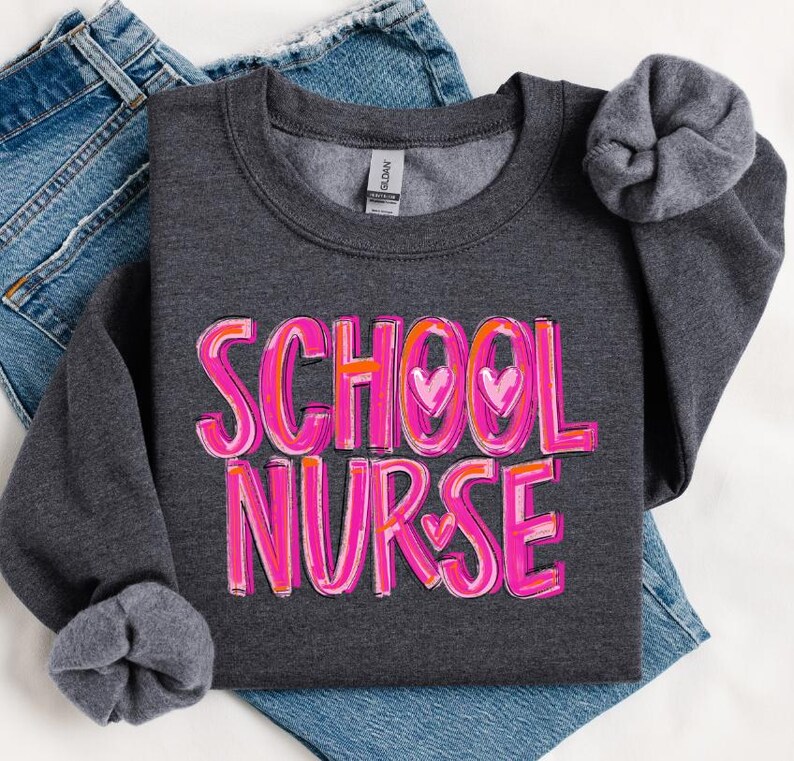 May include: A dark gray sweatshirt with the words "SCHOOL NURSE" in pink and orange lettering with hearts. The sweatshirt is folded and laid on a pair of blue jeans.