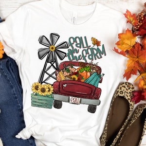 K&ouml;nnte beinhalten: Ein wei&szlig;es T-Shirt mit einem roten Lastwagen, der K&uuml;rbisse, K&uuml;rbisse und Sonnenblumen transportiert. Der LKW hat ein wei&szlig;es Herz an der Seite und ein Nummernschild mit der Aufschrift "Fall". Der Text "Fall on the farm" ist in T&uuml;rkis und Wei&szlig; &uuml;ber dem LKW geschrieben. Im Hintergrund befindet sich eine Windm&uuml;hle.