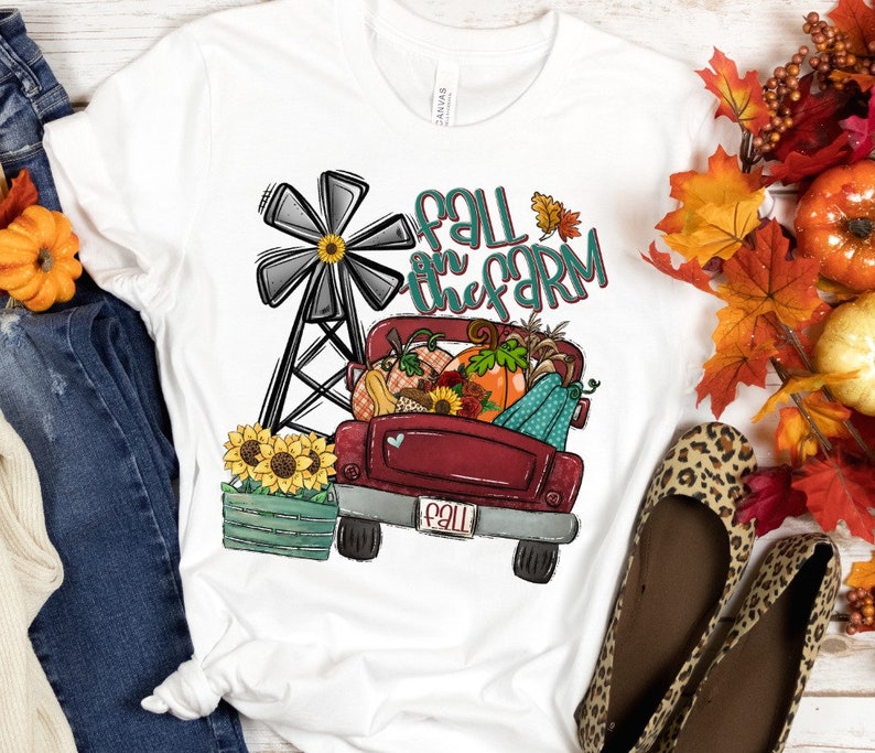 K&ouml;nnte beinhalten: Ein wei&szlig;es T-Shirt mit einem roten Lastwagen, der K&uuml;rbisse, K&uuml;rbisse und Sonnenblumen transportiert. Der LKW hat ein wei&szlig;es Herz an der Seite und ein Nummernschild mit der Aufschrift "Fall". Der Text "Fall on the farm" ist in T&uuml;rkis und Wei&szlig; &uuml;ber dem LKW geschrieben. Im Hintergrund befindet sich eine Windm&uuml;hle.