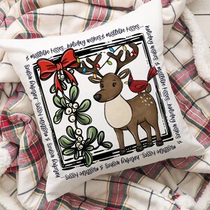 Puede incluir: Un coj&iacute;n cuadrado blanco con un dise&ntilde;o festivo. Presenta un reno de dibujos animados con luces navide&ntilde;as en sus cuernos y un cardenal rojo. El dise&ntilde;o incluye un lazo rojo, mu&eacute;rdago y el texto "holiday wishes & mistletoe kisses".