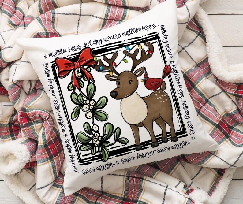 Puede incluir: Un coj&iacute;n cuadrado blanco con un dise&ntilde;o festivo. Presenta un reno de dibujos animados con luces navide&ntilde;as en sus cuernos y un cardenal rojo. El dise&ntilde;o incluye un lazo rojo, mu&eacute;rdago y el texto "holiday wishes & mistletoe kisses".