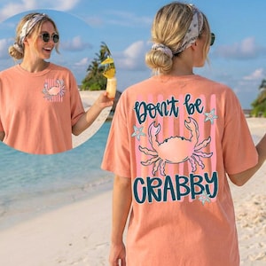 May include: Coral-colored t-shirt with a crab graphic and the text "Don't be Crabby." The shirt features a pocket with a crab design. The design includes pink stripes, teal stars, and teal lettering. The shirt is a casual beachwear item.