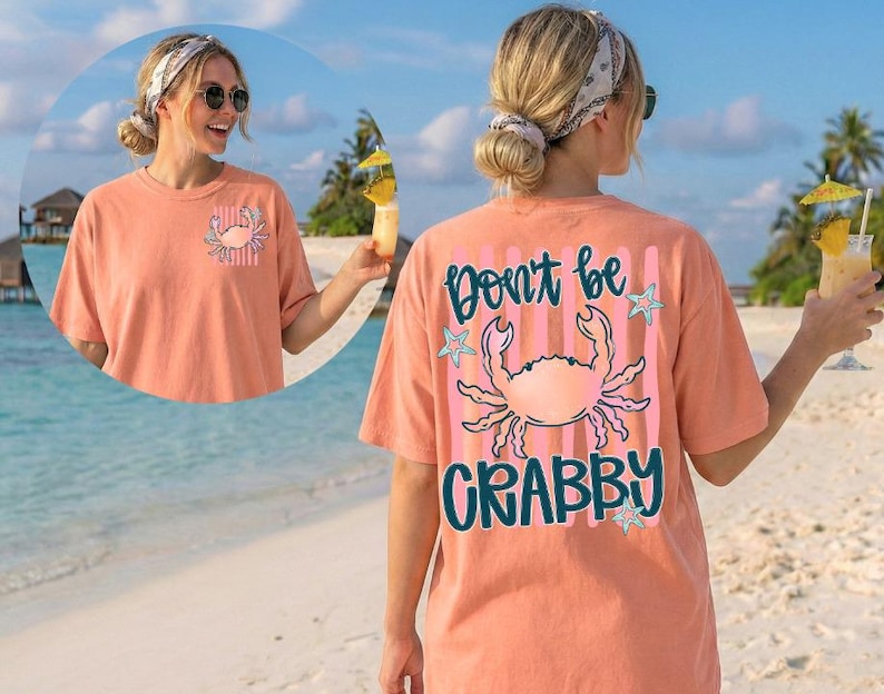 May include: Coral-colored t-shirt with a crab graphic and the text "Don't be Crabby." The shirt features a pocket with a crab design. The design includes pink stripes, teal stars, and teal lettering. The shirt is a casual beachwear item.